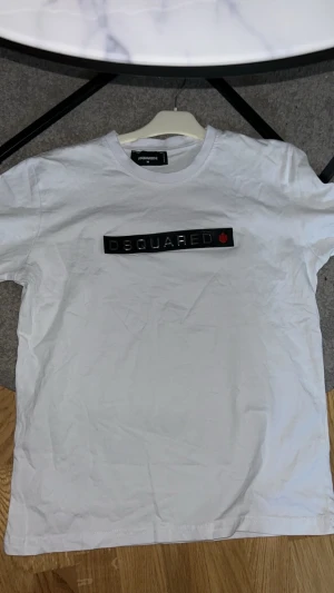 Vit Dsquared2 t-shirt med logga - Säljer en vit t-shirt från Dsquared2 i storlek M. T-shirten har en klassisk passform, rund hals och korta ärmar. På bröstet finns en svart logga med Dsquared2-text och ett rött lönnlöv. Tillverkad i mjuk bomull för skön känsla.
