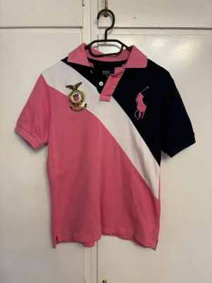 Rosa och marinblå piké från Ralph Lauren - Snygg pikétröja från Ralph Lauren i rosa, marinblått och vitt med klassisk krage och två knappar. Stor rosa logga på bröstet och broderat emblem. Skön bomullskvalitet och korta ärmar. Tröjan är i storlek 164 men passar en S och M. 