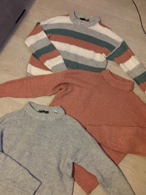 Stickade tröjor i olika färger Lager 157 - Tre stickade tröjor med hög hals. En är randig i grönt, vitt och rost, en är enfärgat rostrosa och en är ljusgrå. Alla har en relaxed fit och ribbade muddar. Perfekta för lager på lager och riktigt mysiga under kalla dagar.