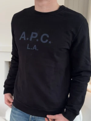 Svart sweatshirt från A.P.C. - Svart sweatshirt från A.P.C. med trycket 'A.P.C. L.A.' i mörkblått framtill. Tröjan har rund halsringning, långa ärmar och ribbade muddar vid ärmslut och nederkant. Perfekt för en avslappnad och stilren look.