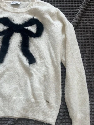 Vit stickad tröja med svart rosett - Supersöt vit stickad tröja från Sweet Sktbs med en stor svart fluffig rosett på bröstet. Tröjan har rund halsringning, långa ärmar och ribbade muddar. Perfekt för dig som gillar trendiga detaljer och mjuka material.