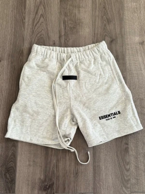 🩶Grå Essentials shorts med snörning🩶 - Snygga ljusgrå shorts från Essentials med svart logga på vänster ben. De har elastisk midja med lång vit snörning och är tillverkade i mjuk bomull. Perfekta för chill dagar eller när du vill ha en clean och enkel look.
