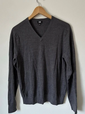 Mörkgrå V-ringad tröja från Uniqlo - Säljer en stilren mörkgrå stickad tröja från Uniqlo med V-ringning. Tröjan har långa ärmar och ribbade muddar vid ärmslut och nederkant. Perfekt att bära över en skjorta eller t-shirt för en clean look.