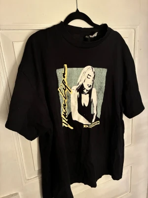 Svart t-shirt med tryck på Dua Lipa från H&M - Cool svart t-shirt från H&M med stort grafiskt tryck framtill i vitt, gult och blått på Dua Lipa. Klassisk rund hals och korta ärmar. Perfekt för dig som gillar statement-plagg och streetstyle.