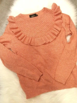 Snygg stickad tröja med volang från ONLY - Superfin stickad tröja i persikorosa från ONLY. Tröjan har lång ärm, rund halsringning med spetsstickat mönster och en bred volangdetalj runt halsen. Perfekt för dig som gillar söta detaljer och mjuka material. Den är i fint använt skick, något nopprig. Storlek XS. 