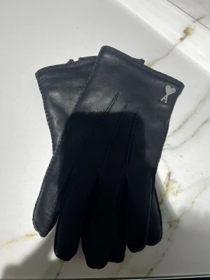 Svarta skinnhandskar med hjärtdetalj - Stilrena svarta handskar i skinn från AMI Paris med en liten silverfärgad hjärtdetalj och bokstaven A på ovansidan. Handskarna har klassisk form och diskreta sömmar på ovansidan. Perfekta för dig som vill ha en clean look.