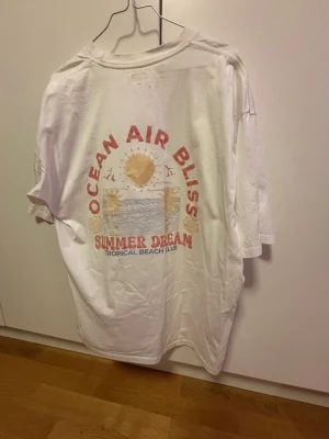 Vit t-shirt med strandtryck over size - Vit t-shirt med färgglatt tryck på ryggen som föreställer en solnedgång, hav och palmer. Texten 'OCEAN AIR BLISS' och 'SUMMER DREAM TROPICAL BEACH CLUB' i rött och blått. Skön och avslappnad passform, perfekt för sommaren.