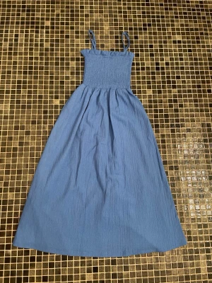 Blå långklänning med smockliv - Säljer en blå långklänning med smala axelband och smockat liv. Klänningen har en enkel, stilren design och är gjord i ett mjukt tyg som faller snyggt. Perfekt för varma dagar och kan stylas på många sätt.