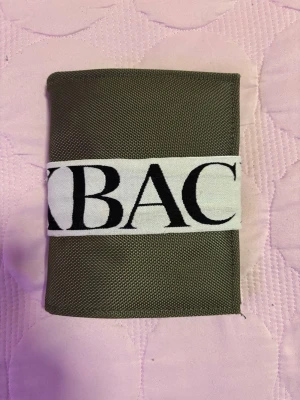 Olivgrön pass plånbok från BACK - Snygg olivgrön plånbok från BACK i slitstarkt tygmaterial. Plånboken har ett vitt band med stor svart logga, flera fack för kort och pass samt en dragkedjeficka. Perfekt för dig som vill ha ordning på allt i en stilren och praktisk design.