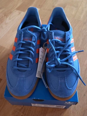 Blå Adidas sneakers med orange detaljer - Säljer ett par blå Adidas sneakers med klassiska orangea stripes och hälparti. Skorna har ovandel i mocka och mesh, samt gummisula i brunt. Snygg retrostil och ikonisk design som sticker ut. Perfekta för dig som gillar färg och streetwear.