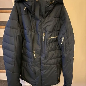 Mörkblå skidjacka från Peak Performance - Snygg mörkblå skidjacka från Peak Performance med huva och flera dragkedjeförsedda fickor framtill. Jackan har en quiltad design och logga på bröstet. Perfekt för kalla vinterdagar och håller dig varm med sin tjocka vaddering.