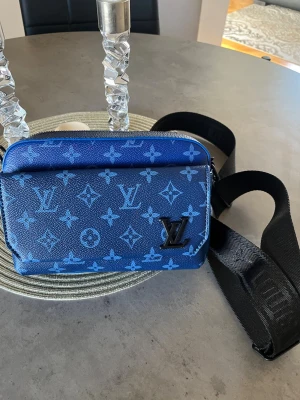 Blå Louis Vuitton axelväska - Snygg blå axelväska från Louis Vuitton med ikoniskt monogrammönster i ljusblått. Väskan har en justerbar svart axelrem, dragkedja upptill och en praktisk ytterficka. Tillverkad i tåligt canvasmaterial med metallogo framtill. Perfekt för dig som vill ha en trendig och exklusiv accessoar.