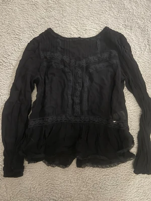 Svart blus med spets från Gina Tricot Stockholmsstil  - Snygg svart blus från Gina Tricot i transparent material med spetsdetaljer och volang nedtill. Blusen har rund halsringning och långa ärmar. Perfekt för dig som gillar en romantisk och trendig stil.