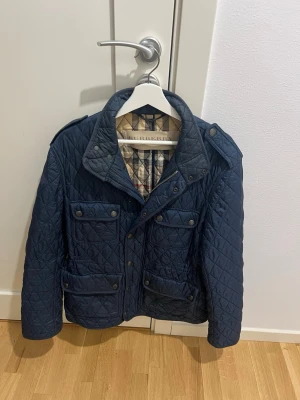 Burberry Brit Quilted Field Jacket – Klassisk Marinblå (Bra skick!) - Säljer nu min klassiska diamantquiltade field jacket från Burberry Brit. Jackan är i en tidlös mörkblå (navy) färg med det ikoniska rutiga fodret (Nova Check) som syns tydligt när man viker upp kragen eller har jackan öppen. Detta är en perfekt jacka för vår och höst som kombinerar brittisk elegans med praktisk funktion. Små defekter se bild 