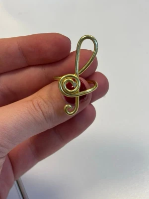 Guldfärgad ring formad som en G-klav - Ring i guldfärg formad som en G-klav. Ringen består av gulfärgad metalltråd med en kreativ formning. Passar för dig som gillar unikat gjorda smycken. 