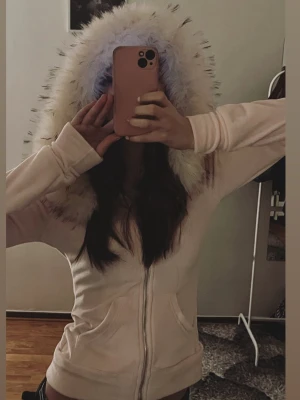 Ljusrosa hoodie med fuskpäls - Säljer en ljusrosa hoodie med dragkedja och stor huva kantad med fluffig fuskpäls. Hoodien har fickor framtill och är gjord i mjukt sweatshirtmaterial. Perfekt för dig som vill ha en cozy och stylish look.
