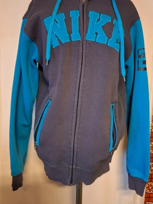 Nike hoodie med dragkedja, blå ärmar och huva - Snygg hoodie från Nike i mörkgrått med turkosa ärmar och detaljer. Stor logga framtill och dragkedja hela vägen. Två fickor med dragkedja och huva med matchande snören. Perfekt för dig som gillar sportig och avslappnad stil.