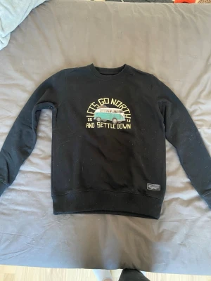 Svart sweatshirt från SQRTN Company - Svart sweatshirt från SQRTN Company med tryck av en grön minibuss och texten 'Let's go north and settle down'. Tröjan har rund halsringning, ribbade muddar och är tillverkad i mjukt material. Storlek XXS men sitter som en XS