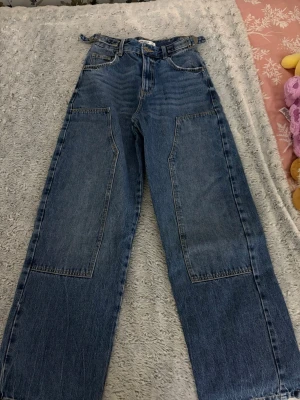 Blåa wide leg jeans  - Säljer ett par blåa wide leg jeans från Pull&Bear med stora fickor framtill och justerbar midja. Jeansen har hög midja och raka ben, perfekta för en avslappnad och trendig look. Klassisk denim med kontrastsömmar och bälteshällor.
