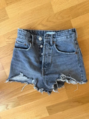 Högmidjade jeansshorts med råa kanter - Snygga högmidjade jeansshorts i blå denim med råa, fransiga kanter och slitna detaljer. Klassisk femficksmodell med knappar framtill och bälteshällor. Perfekta för en avslappnad och trendig sommarstil. 