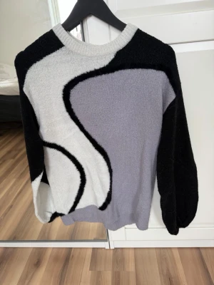 Stickad tröja med abstrakt mönster - Säljer en stickad tröja med coolt abstrakt mönster i svart, grått och vitt. Tröjan har rund halsringning och långa ärmar. Materialet känns mjukt och mysigt, perfekt för kyligare dagar. Passar dig som gillar unik och trendig stil.