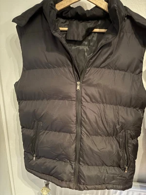 Svart dunväst med huva - Stilren svart dunväst med hög krage och två praktiska fickor med dragkedja. Västen har quiltad design och stängs med en rejäl dragkedja framtill. Perfekt att slänga över en hoodie för en casual look. Materialet är glansigt och ger en modern känsla.