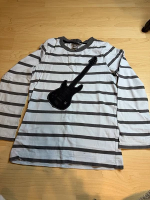 Randig långärmad topp H&M - Säljer en vit och grå randig långärmad topp från H&M med ett coolt svart gitarrtryck på bröstet. Perfekt för dig som gillar musik och vill sticka ut lite extra. Tillverkad i mjuk bomull för skön känsla hela dagen.
