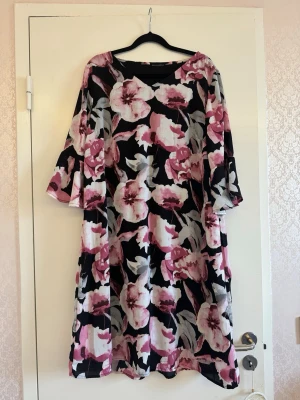 Blommig svart och rosa klänning - En loose-fit klänning med blommigt mönster i rosa, vitt och grått på svart botten. Klänningen har trekvartsärmar och rund halsringning. Perfekt för dig som gillar färg och mönster. Storlek L. Bröstmåttet är ca 100cm (50cm x 2). 