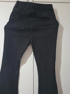 Svarta bootcut jeans med rå kant - Säljer ett par svarta bootcut jeans med hög midja och fem knappar framtill. Jeansen har råa, fransiga benslut och klassiska bakfickor. Materialet är stretchigt denim som sitter snyggt och bekvämt. Perfekta för en trendig och avslappnad look.