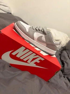 Nike Dunk Low grå/vit sneakers - Nike Dunk Low sneakers i stilren grå och vit färgkombination. Skon har klassisk låg siluett, vita paneler i läder och ljusgrå overlays samt matchande grå snören och sula. Perfekt för dig som gillar clean och tidlös streetstil.