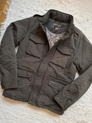 Vadderad Fieldjacket - Woolrich  - En extremt kvalitativ jacka som nästan har en vaxad feel utanpå. Varmt foder, man kommer inte att gå kall med denna. Nedersta knappen går ej att knappa vilket inte påverkar användningen (Bild 4). Hör av dig vid frågor!