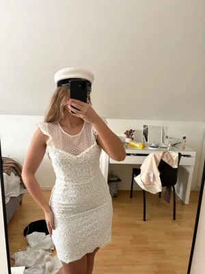 Vit spetsklänning  Studentklänning - Säljer en superfin vit kort spetsklänning med volangdetaljer vid axlarna och transparent mesh upptill. Klänningen har figurnära passform och v-ringad illusion vid halsen. Perfekt till student eller sommarens alla tillfällen! Användes aldrig till studenten köpte en annan.