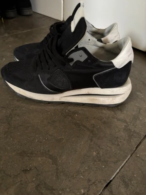 Svarta Philippe Model sneakers - Snygga svarta sneakers från Philippe Model med vita detaljer och grå inslag. Ovandelen är i en mix av mocka och mesh för både stil och komfort. Klassisk låg modell med svart snörning och vit sula som ger en sportig vibe. Storlek 40 men passar 41 ev också 42. Nypris 3000