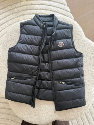 Svart tunn dunväst från Moncler storlek 164 - Snygg svart dunväst från Moncler med quiltad design och hög krage. Västen har två dragkedjeförsedda fickor framtill och Moncler-logga på bröstet. Perfekt för lager-på-lager och ger en clean look till din outfit. Dragkedjan har gått sönder nertill men går att byta/fixa. 1500kr 