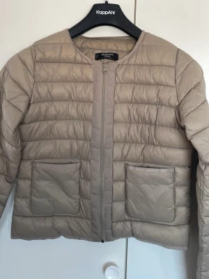 Beige pufferjacka från Mango - Stilren beige pufferjacka från Mango med rund halsringning och två stora framfickor. Jackan har dragkedja framtill och quiltad design med horisontella sömmar. Perfekt för kalla dagar och enkel att matcha till olika outfits.