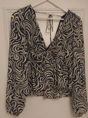 Svartvit mönstrad blus med v-ringning - Trendig blus med svartvitt abstrakt mönster, djup v-ringning och knytband i nacken. Långa puffärmar med resår vid ärmslut och volang nedtill. Tunn och luftig i materialet, perfekt för att sticka ut i mängden.