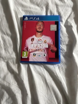 FIFA 20 till PS4 - FIFA 20 till PlayStation 4 med blå plastfodral. Omslaget har en röd bakgrund och visar en fotbollsspelare i vit Real Madrid-tröja. Perfekt för dig som gillar fotbollsspel och vill utmana vänner i klassiska matcher.