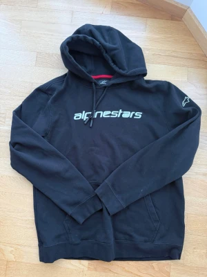 Svart Alpinestars hoodie med tryck - Snygg svart hoodie från Alpinestars med stor logga på bröstet och ikonisk stjärna på ärmen. Klassisk känguruficka framtill, justerbar huva med snören och mjukt material som känns skönt mot huden. Perfekt för dig som gillar streetwear och motorsport vibes.