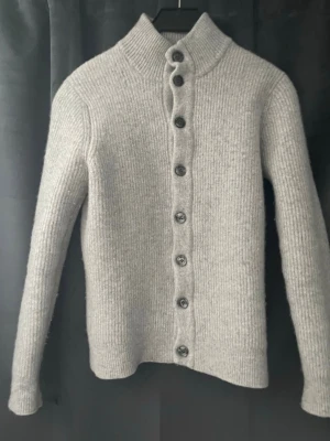 Grå stickad kofta från Massimo Dutti - Säljer en stilren grå stickad kofta från Massimo Dutti i ull och kashmir. Koftan har hög krage, långa ärmar och knäppning med åtta mörka knappar framtill. Perfekt för lager på lager och en clean look. Vid snabbköp kan jag tänka sänka priset till 600kr.