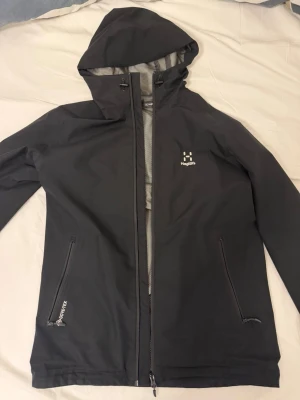 Haglöfs jacka - Snygg svart regnjacka från Haglöfs med huva och dragkedja framtill. Jackan har två fickor med dragkedja och är tillverkad i GORE-TEX-material som andas. Insidan har meshfoder för extra komfort. Perfekt för blöta dagar när du vill hålla dig torr.