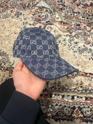 Blå denimkeps från Gucci - Snygg blå keps från Gucci i denim med det klassiska GG-monogrammet över hela kepsen. Böjd skärm och justerbar baksida. Perfekt för dig som vill ha en trendig accessoar med lyxig känsla och streetwear-vibe.