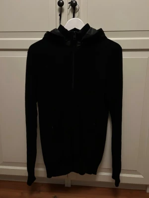 Svart cardigan  - Säljer en svart cardigan  två fickor med dragkedja. Cardiganen har en klassisk huva och är tillverkad i ett stickat material som känns både mjukt och bekvämt. Perfekt för en avslappnad och stilren look.