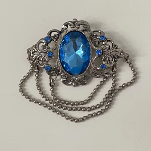 Vintage brosch med blå sten - Snygg vintage brosch i silverfärgad metall med en stor oval blå glassten i mitten och mindre blå stenar runtom. Dekorativt mönster och tre kedjor i metall ger en unik och elegant look. Perfekt accessoar för att lyfta din outfit.