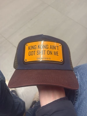 John Hatter & Co keps - Säljer en brun och svart truckerkeps från John Hatter & Co med ett gult metallmärke framtill där det står 'KING KONG AIN'T GOT SHIT ON ME'. Kepsen har mesh på sidorna och justerbar snapback baktill. Perfekt för dig som vill sticka ut med en cool statement-keps.