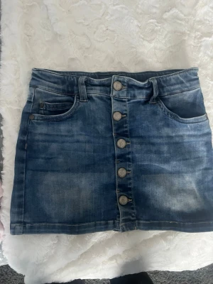 Blå kortkjol med knappar - Snygg blå jeanskjol i kort modell med fem knappar fram. Jättesnygg säljer på grund av att jag tycker den är för kort. funkar säkert för någon i XS med beroende på hur kort man vill ha den. 