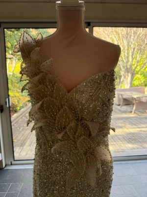 Champagnefärgad långklänning med pärlor - Elegant långklänning i champagnefärg med en axel och stora blommiga applikationer. Klänningen är täckt av pärlor och paljetter som ger ett glittrigt intryck. Materialet ser ut att vara en blandning av siden och polyester. Perfekt för bal eller fest. Köpt för 9000 pris går att diskutera vid snabb affär