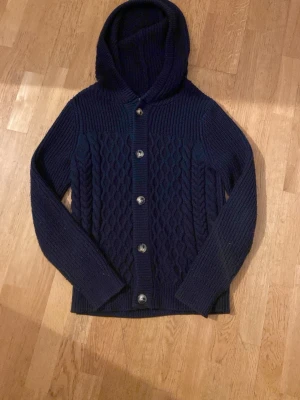 Mörkblå stickad hoodie Pier One - Säljer en mörkblå stickad hoodie från Pier One med snyggt kabelstickat mönster framtill. Tröjan har huva. Storleken är S men passar mer som en M. Riktigt skön och är len på huden!