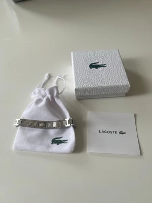 Silverfärgat armband från Lacoste - Snyggt armband i silverfärgad metall från Lacoste med klassisk krokodil-logga på spännet. Armbandet har rektangulära länkar med diskret mönster och kommer i originalförpackning med tygpåse och manual. Perfekt accessoar för en stilren look. Den är cirka 20cm lång men har en länkbortagare som kan göra den till din önskade storlek.