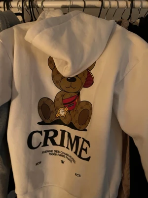 Vit hoodie med nalle och text CRIME - Vit hoodie med stor huva och tryck av en nallebjörn på ryggen. Nallen har en röd keps och magväska, samt mönstrade detaljer. Under motivet står texten CRIME och adress i Paris. Hoodien har en avslappnad passform och är gjord i mjukt material. Liten brönnmärke därför jag säljer den då billigt men den synd knappt 