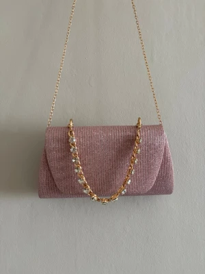 Rosa glittrig kuvertväska med kedja - Snygg rosa kuvertväska med glittrigt tyg och guldfärgad kedja. Väskan har även ett dekorativt handtag med gnistrande stenar som ger extra lyxig känsla. Perfekt att bära över axeln eller i handen för en trendig look.